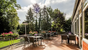 An outdoor patio area with tables and chairs, surrounded by greenery and trees. - Messehotel Köln-Deutz (Cologne)
