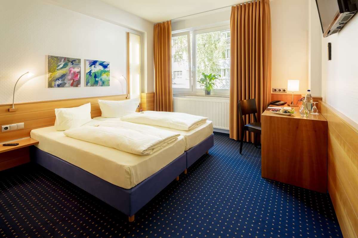 A hotel room with a double bed, a desk with a chair, a small table with a plant, and artwork on the wall.