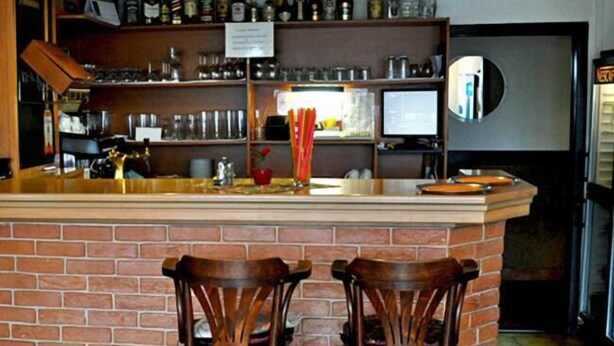 A bar area with a brick counter, two wooden bar stools, shelves with bottles and glasses, and a sign above the counter. - Hotel Milotel (Olomouc)