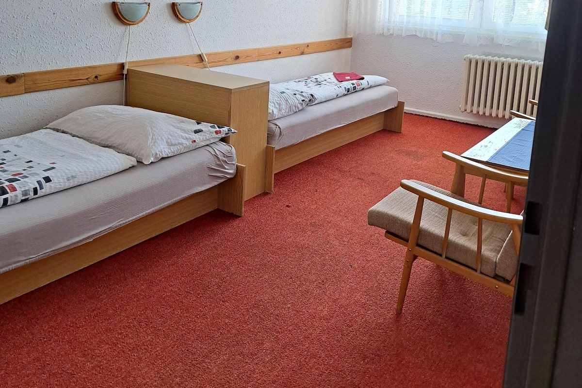 A room with two single beds, a wooden desk, a chair, and a window with sheer curtains.