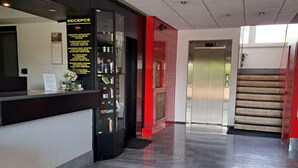 A hotel reception area with a reception desk, a digital clock, and a staircase leading to an elevator. - Hotel Milotel (Olomouc)