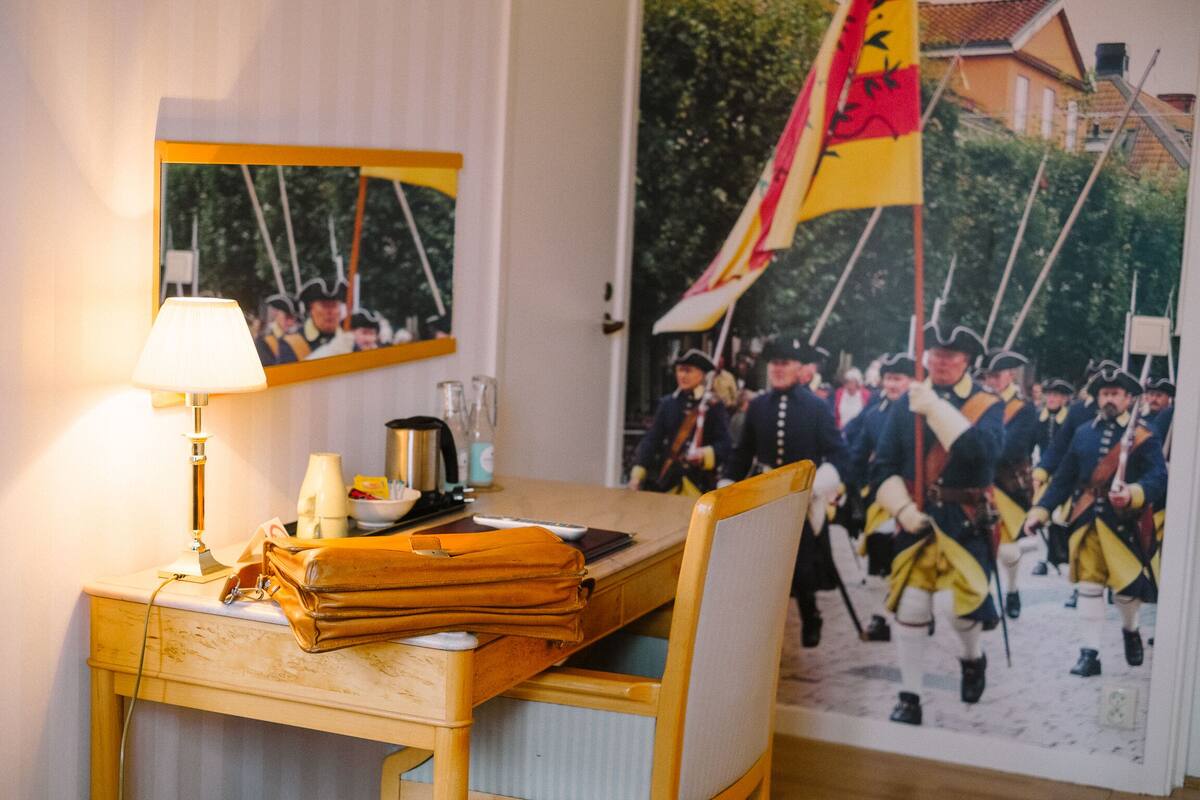 Habitación de hotel con un escritorio de madera, una lámpara, un bolso de mano y un cuadro enmarcado que representa una escena de batalla histórica.