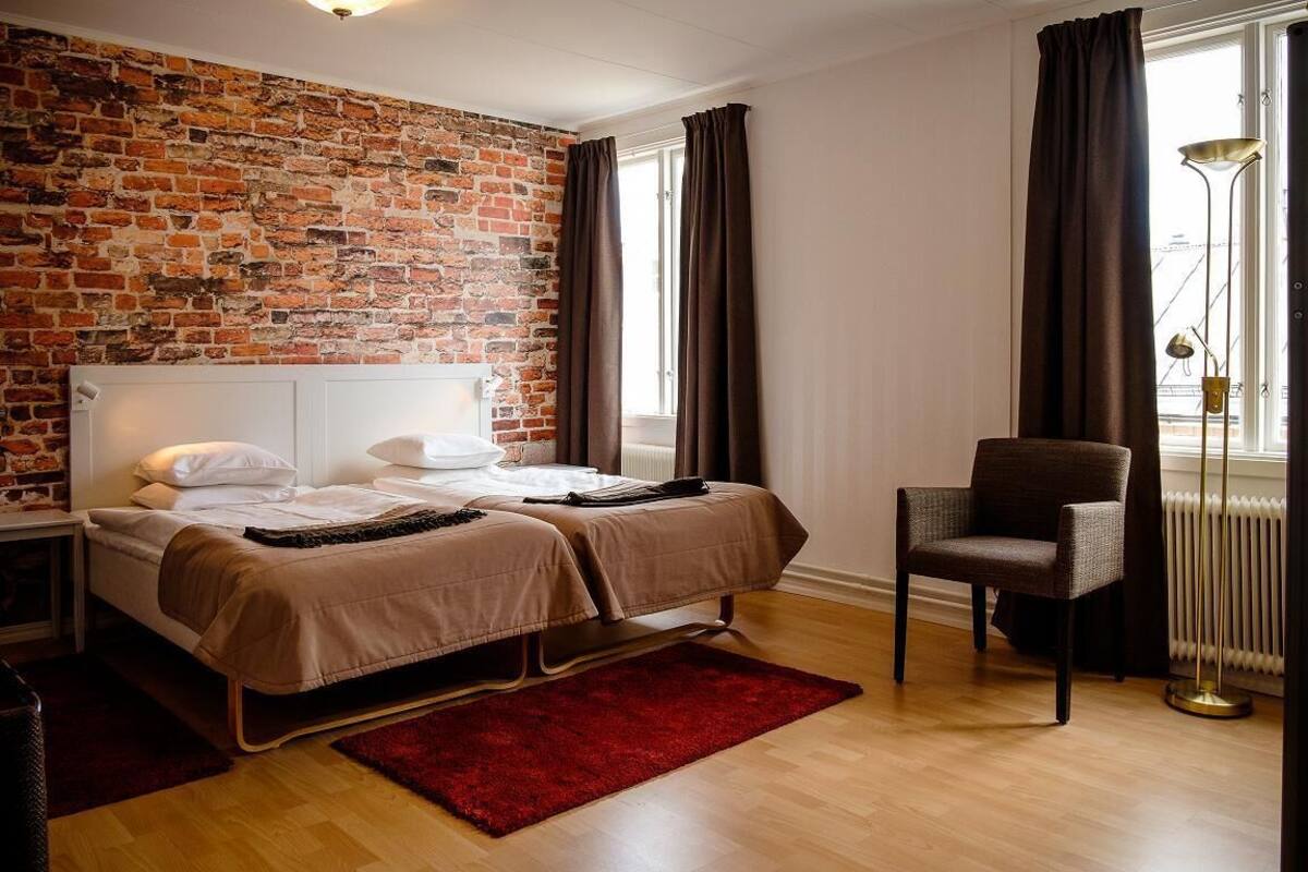 Habitación de hotel con pared de ladrillo, dos camas, una alfombra roja, una silla y una ventana con cortinas.
