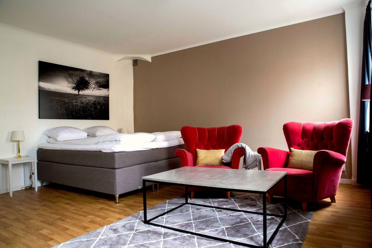 Un dormitorio con una cama, dos sillones rojos, un sofá gris, una mesita y un cuadro en la pared.