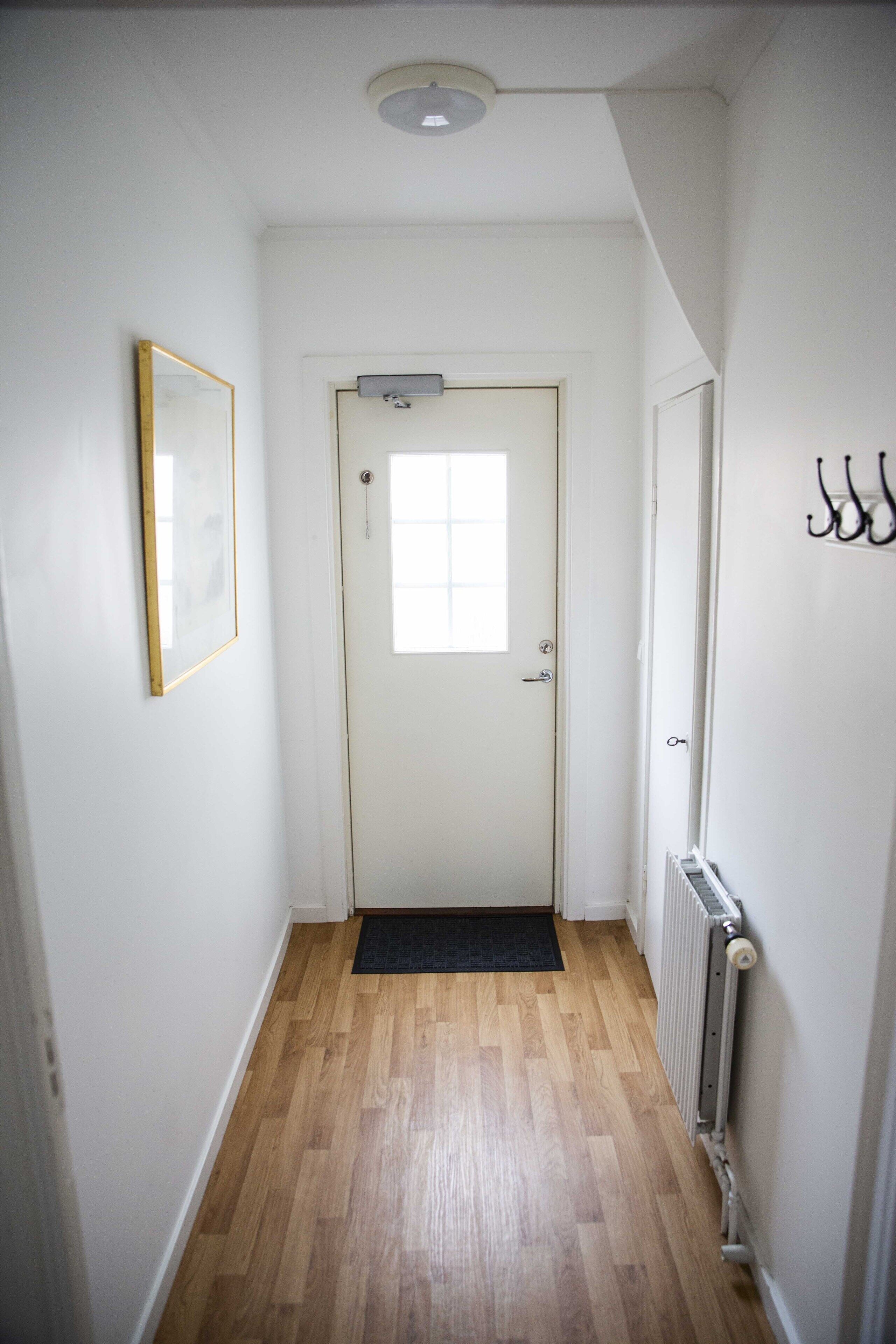A narrow hallway with wooden flooring, a white door with a window, and a radiator on the right.