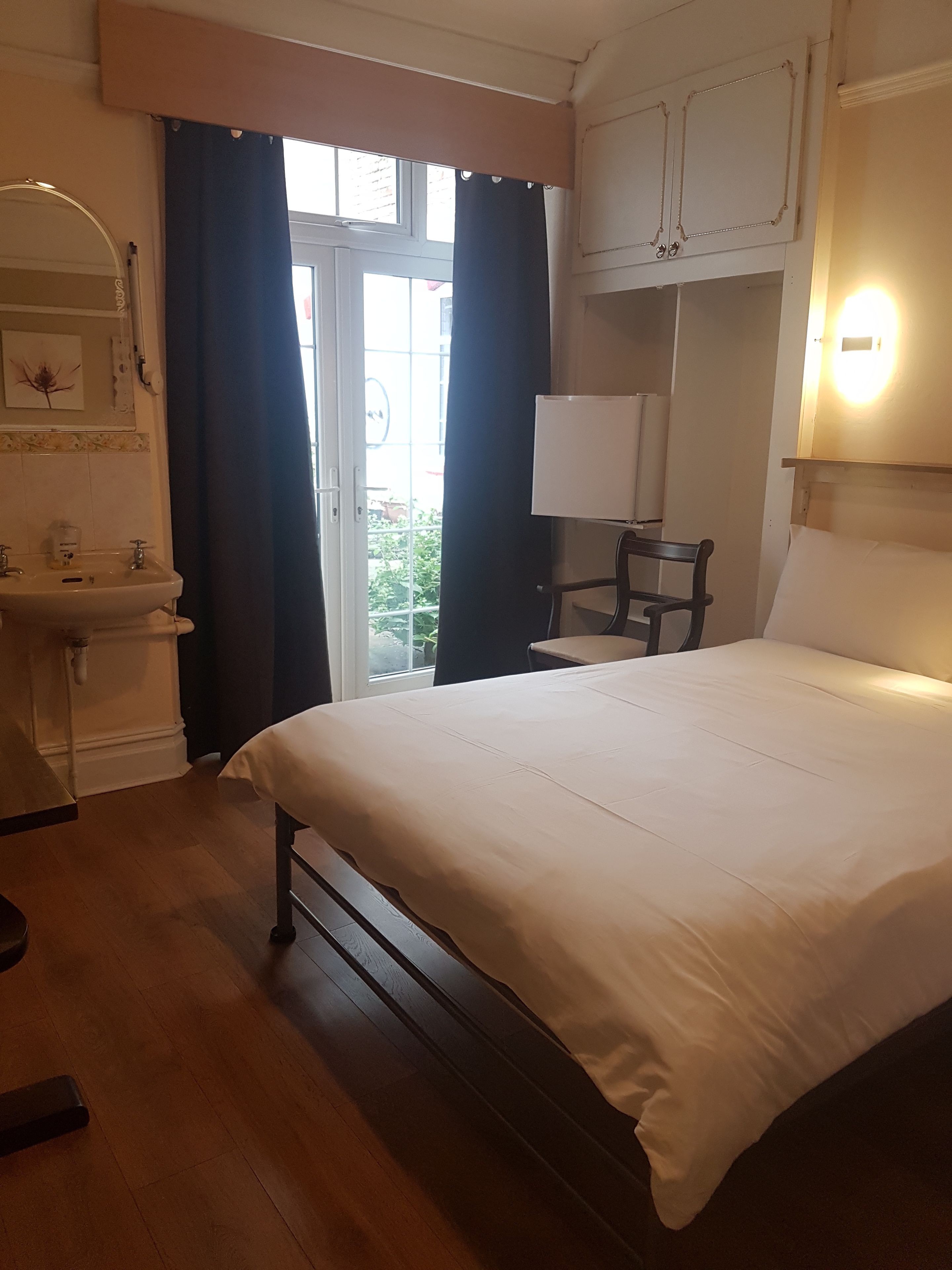 Une chambre d’hôtel comprenant un lit, une table de chevet, une chaise, une fenêtre avec des rideaux, un lavabo et un miroir.