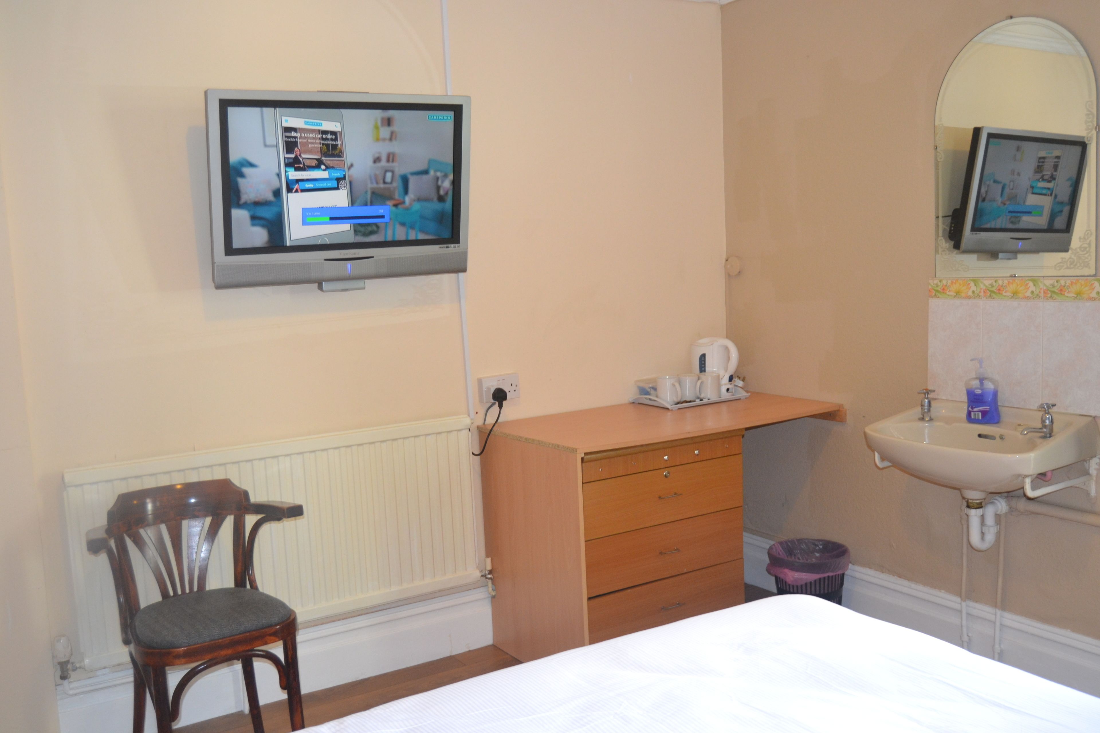 Ein Hotelzimmer mit einem Bett, einer Holzkombination, einem an der Wand befestigten Fernseher, einem kleinen Tisch mit Wasserkocher und einem Badezimmer mit Waschbecken und Spiegel.