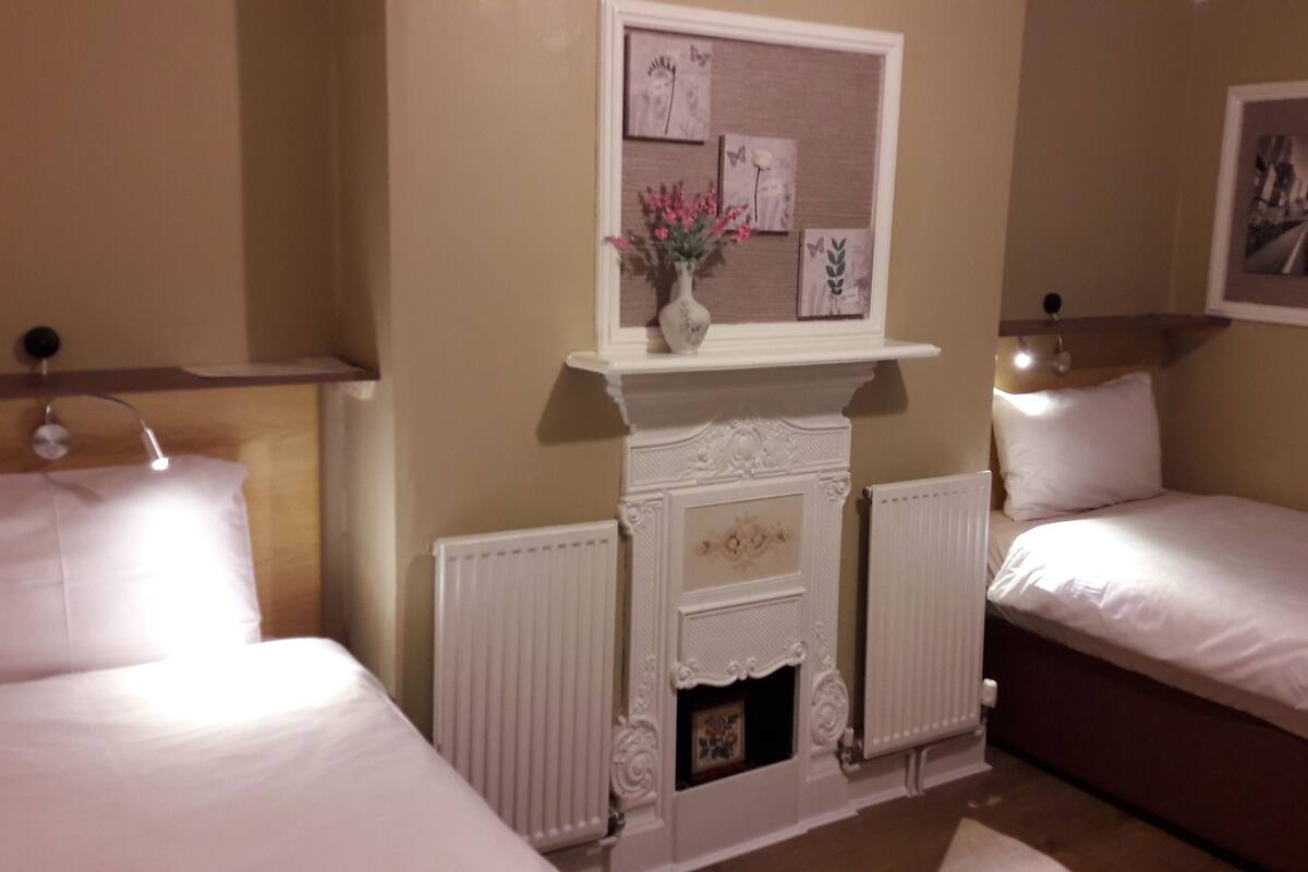 A hotel room with two beds, a white fireplace with a vase of flowers, and a bulletin board with artwork.