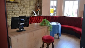 Une pièce avec un banc rembourré de couleur rouge, un bureau en bois avec un écran d’ordinateur, une petite table recouverte d’une nappe bleue et un mur en briques.