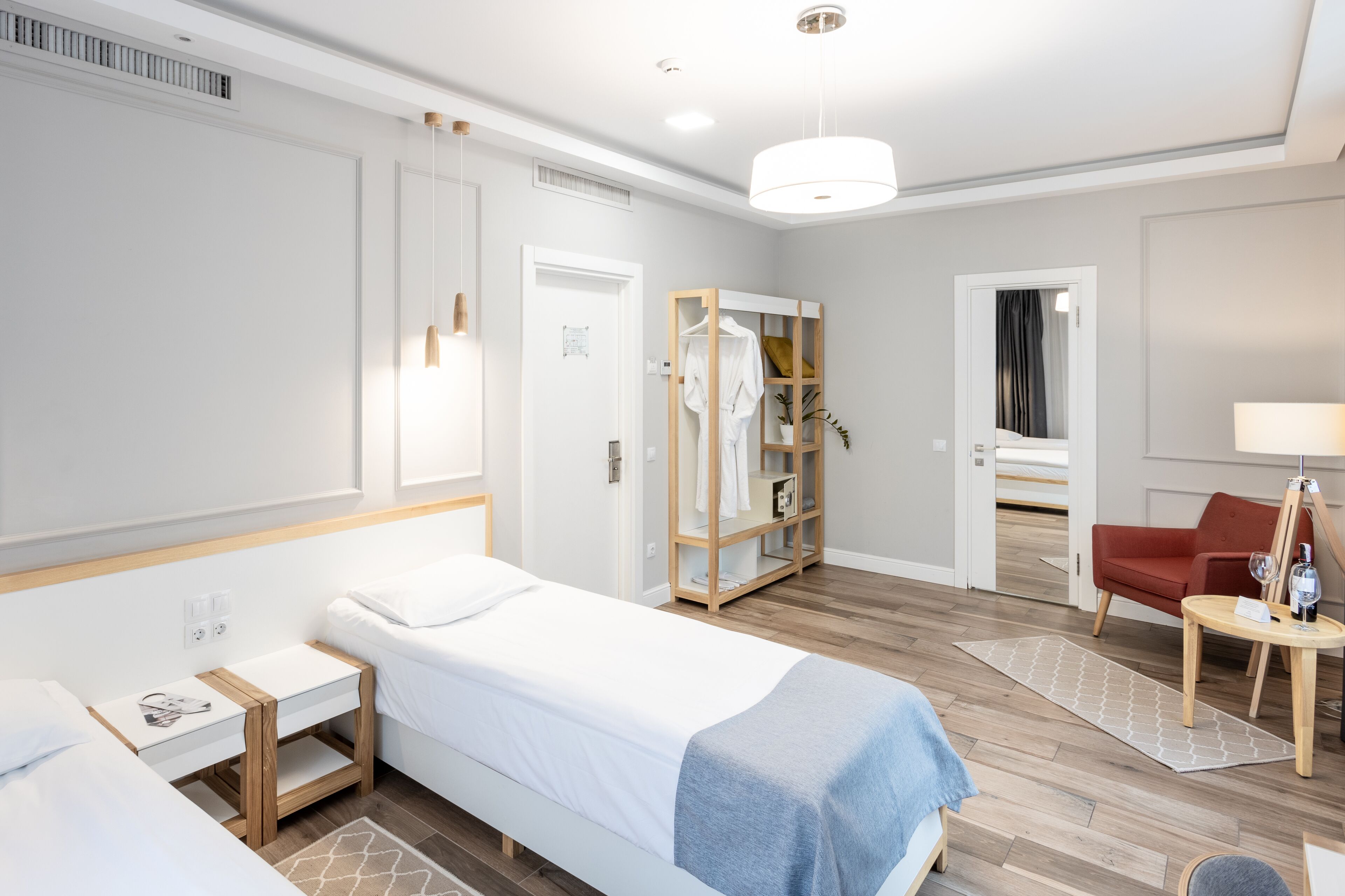 A modern hotel room with a single bed, bedside table, wardrobe, and a small seating area with a chair and table.