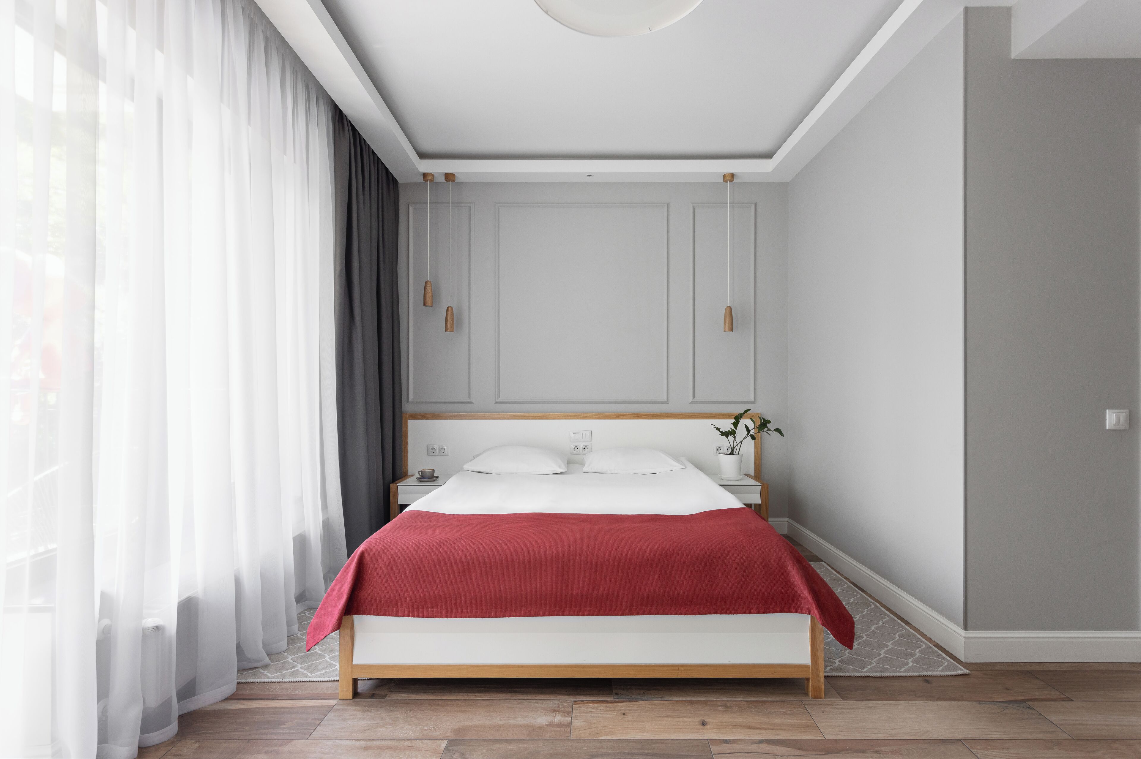Un dormitorio moderno con cama de madera, ropa de cama blanca, un plaid rojo y cortinas traslúcidas.
