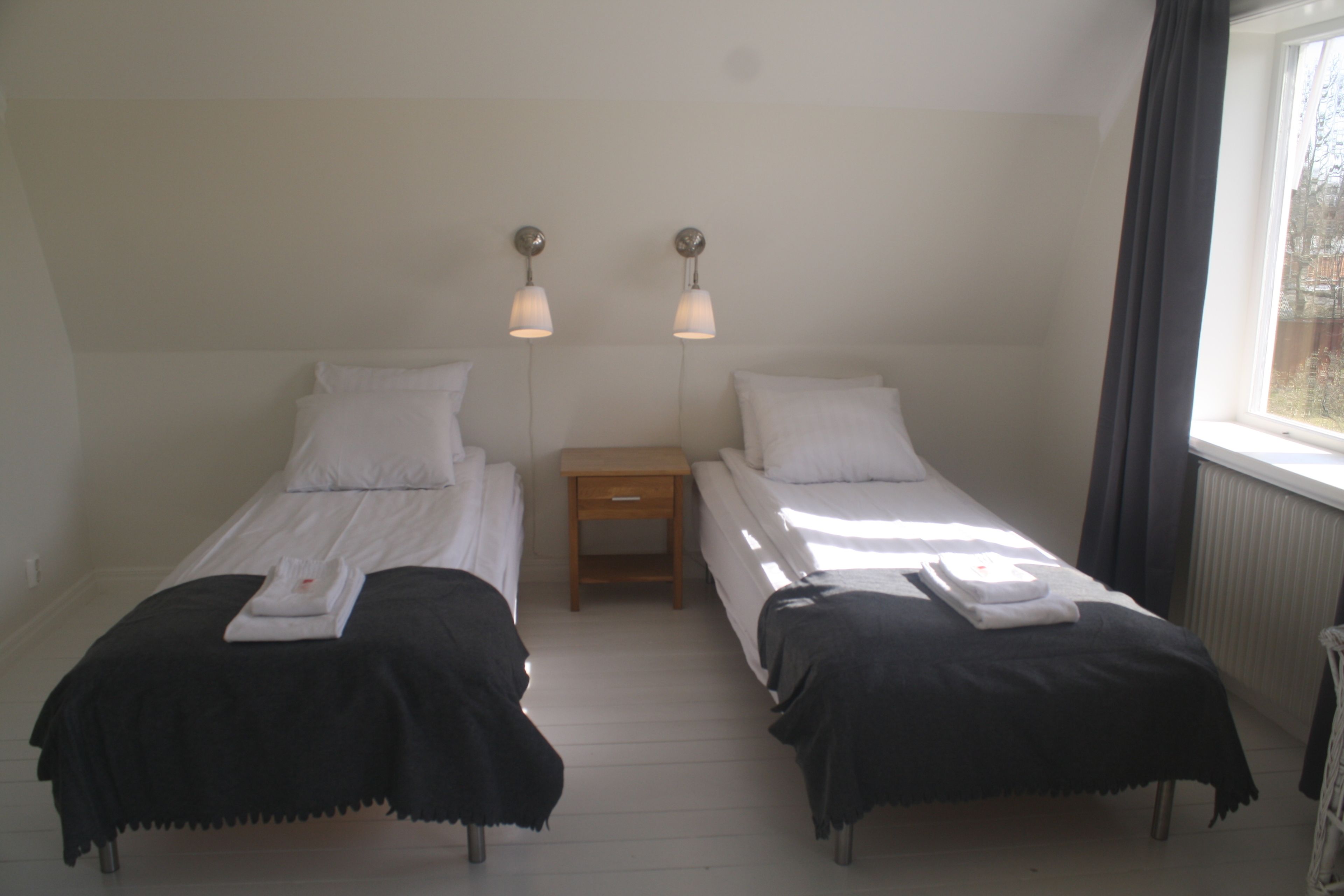 A room with two single beds, each with a white pillow and a black bedspread. There are two bedside tables with lamps, and a window with curtains on the right side.