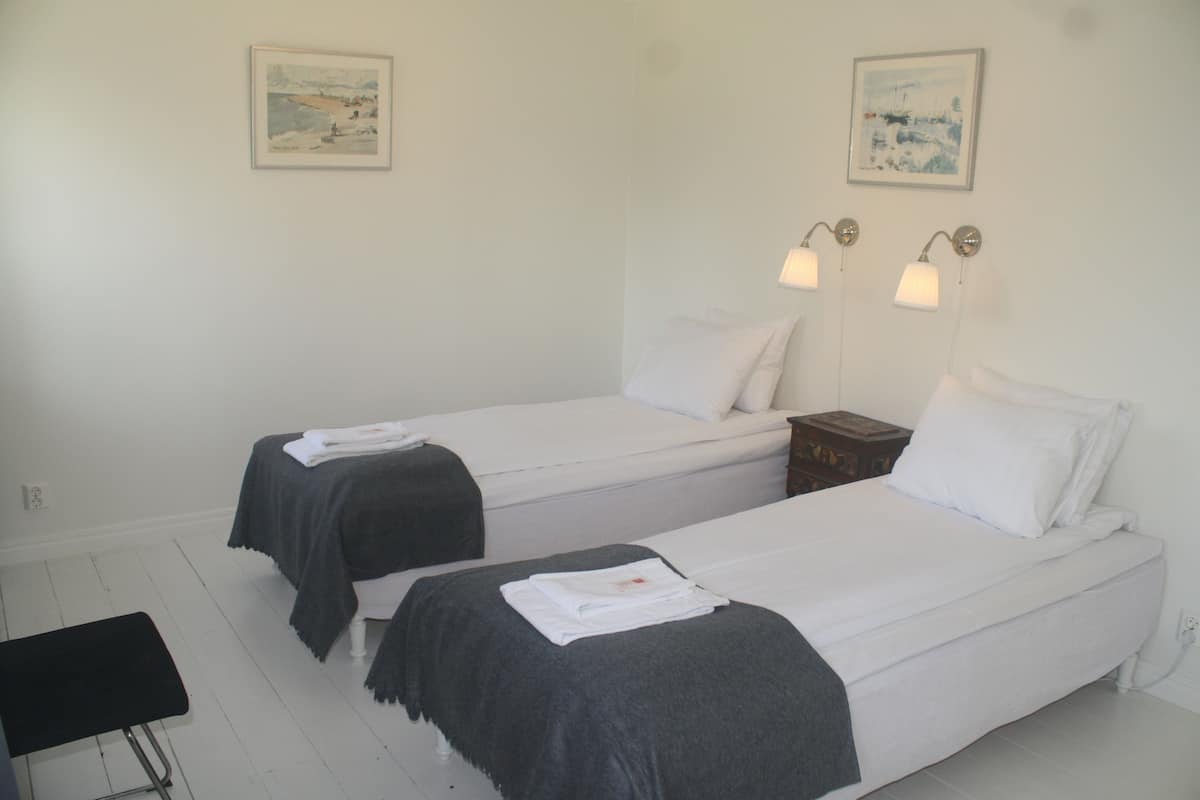 A hotel room with two single beds, each with a white mattress and a dark grey blanket. There are two wall-mounted lamps on either side of the beds.