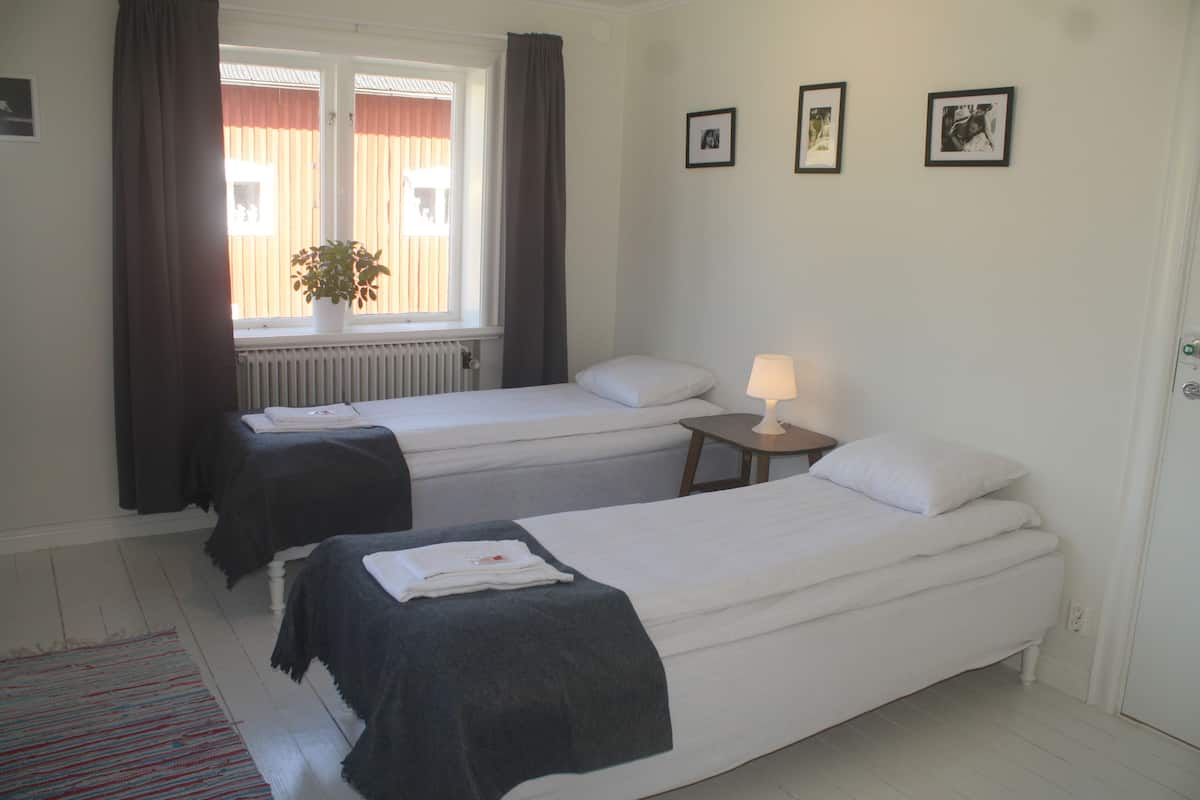 A room with two single beds, a small table with a lamp, a radiator, and a window with curtains.