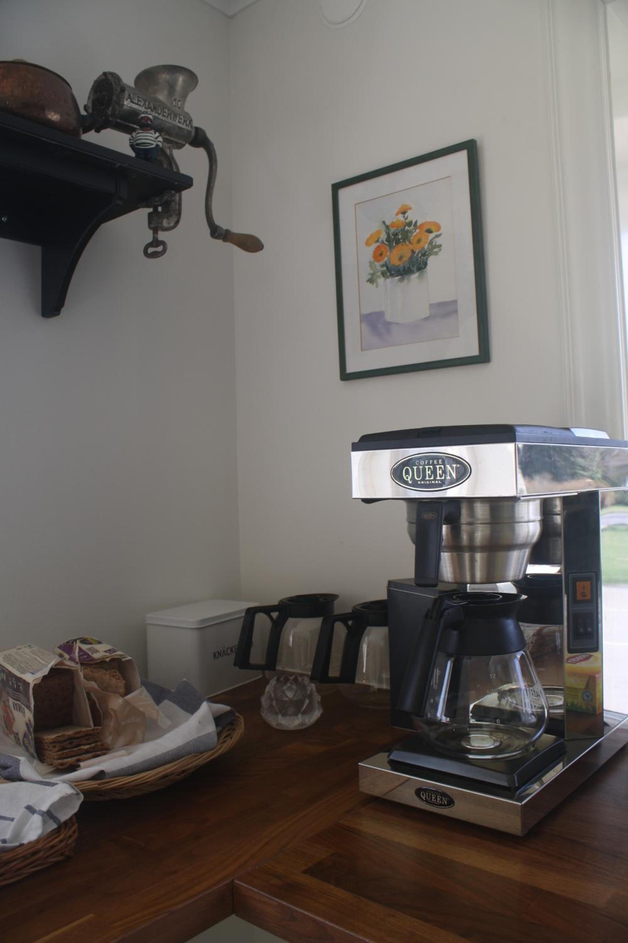 Eine Kaffeemaschine auf einem Holztisch, neben einem Brotkorb und einem gerahmten Bild an der Wand.
