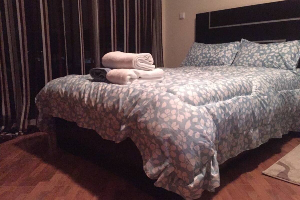 A neatly made bed with a floral-patterned bedspread and pillows, a rolled-up towel, and a dark headboard.