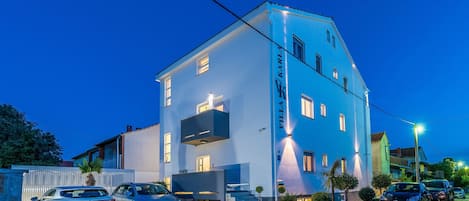 A modern two-story building with a blue facade, a balcony, and a parking area with several cars.