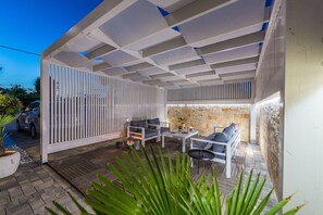 Un patio al aire libre con terraza de madera, mobiliario blanco y pared de piedra.