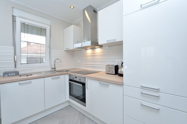 A modern kitchen with white cabinets, a built-in oven, a stainless steel range hood, and a window with blinds.