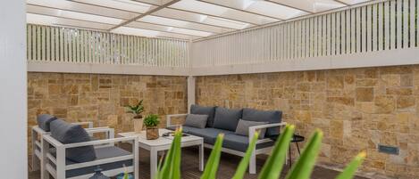 Un patio al aire libre con un muro de piedra, mobiliario blanco y un techo con diseño de cuadrícula.