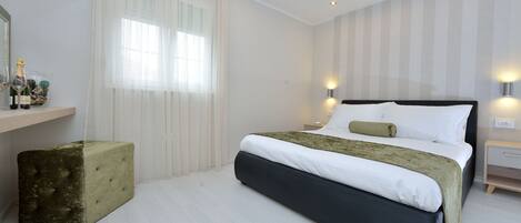 Un dormitorio moderno con una cama grande, una mesita de noche, una pequeña silla y una ventana con cortinas traslúcidas.