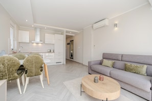 Una sala de estar moderna con un sofá gris, una mesa de centro redonda de madera y un comedor con una mesa blanca y sillas tapizadas de verde.