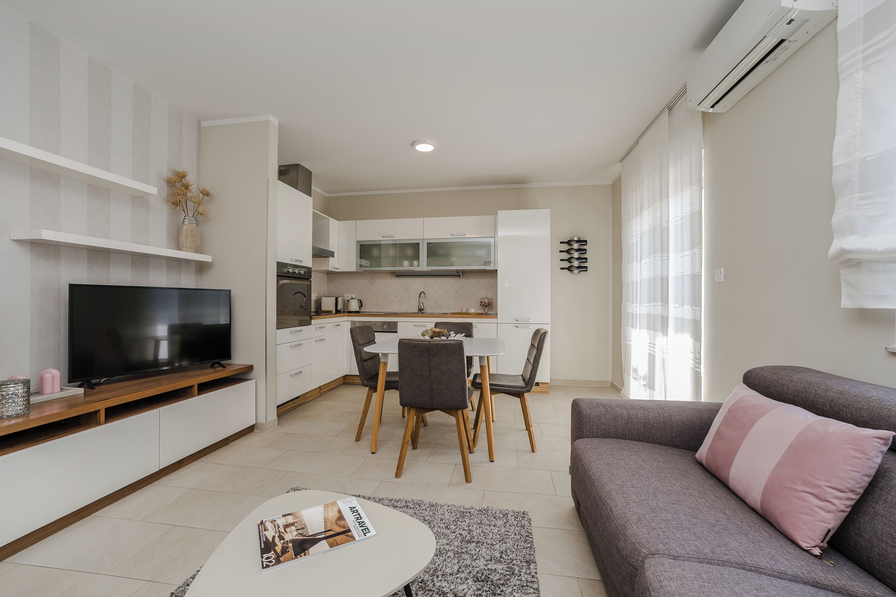 A modern living room with a flat-screen TV, a grey sofa, and a dining area with a table and chairs.