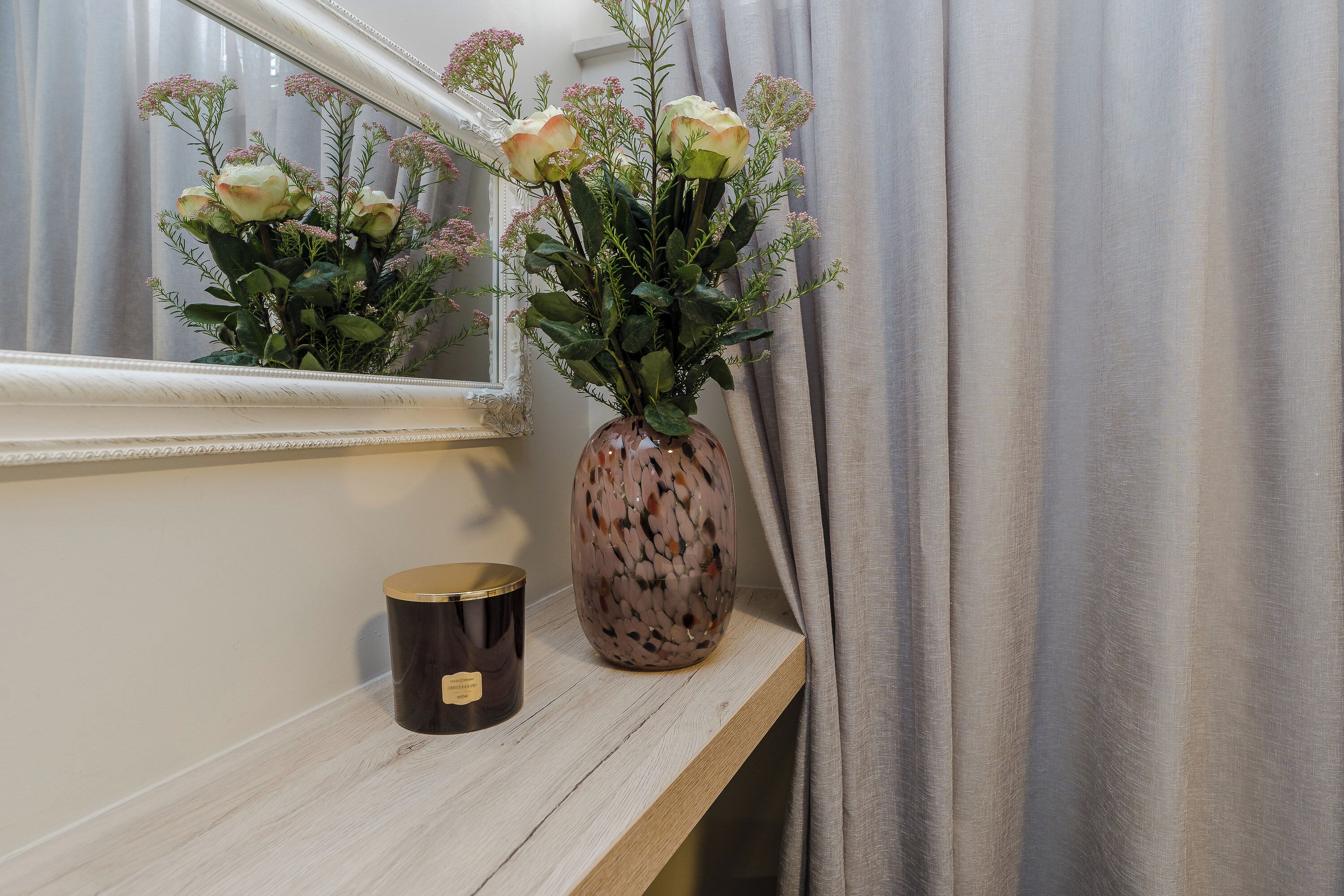 A mirror reflecting a vase with flowers and a candle on a wooden shelf.