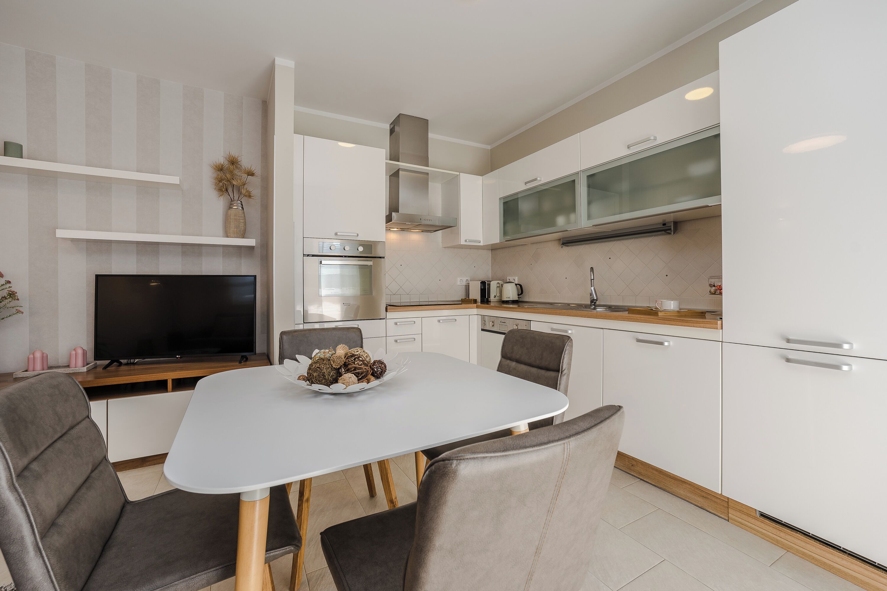 Une cuisine moderne avec des armoires blanches, un four encastré et un coin repas comprenant une table blanche et deux chaises.