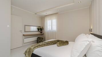 A modern bedroom with a large bed, a bedside table with a mirror, and a window with sheer curtains.