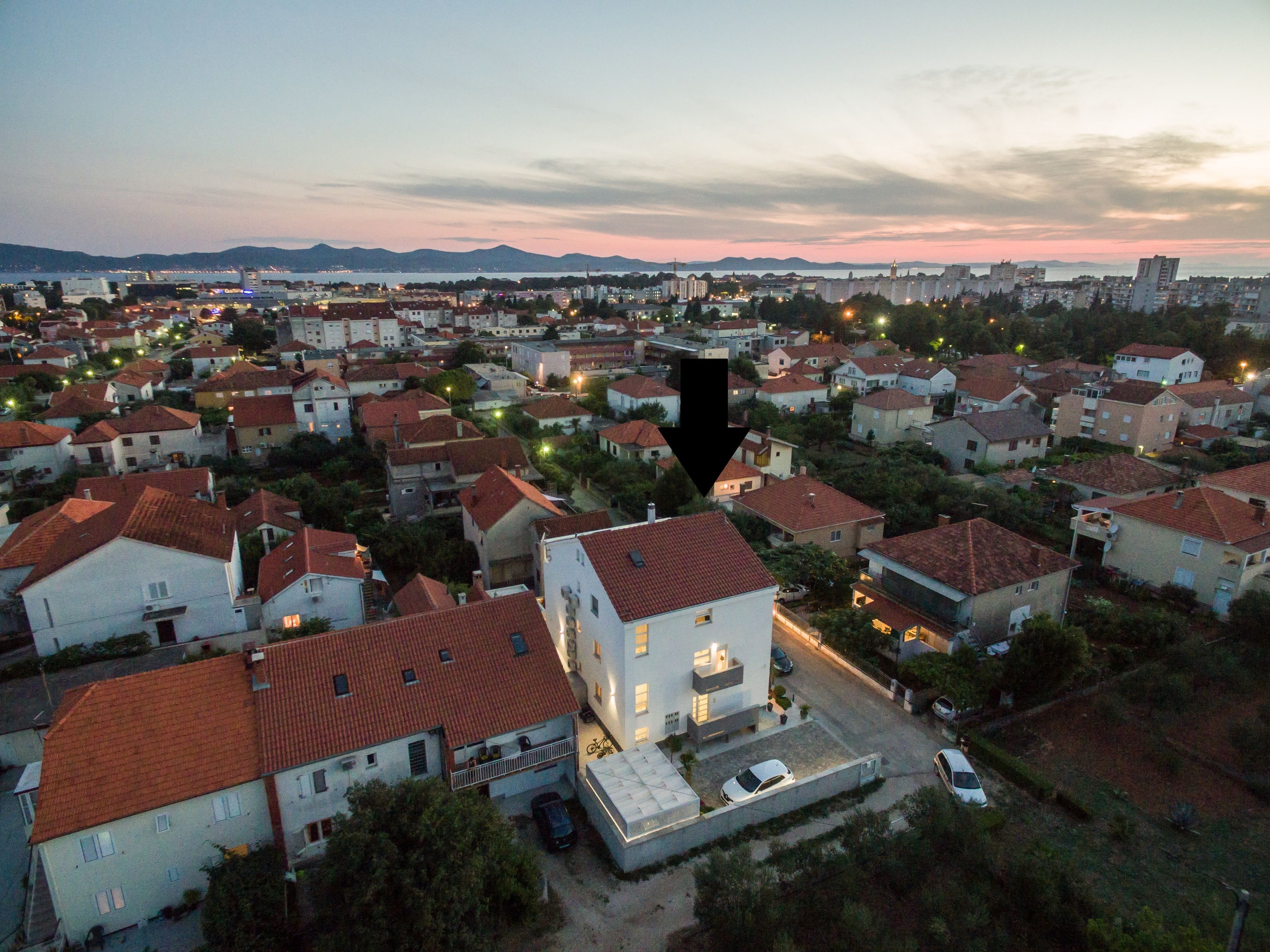 An aerial view of a residential area with houses, a prominent white building with a balcony, and a body of water in the background.