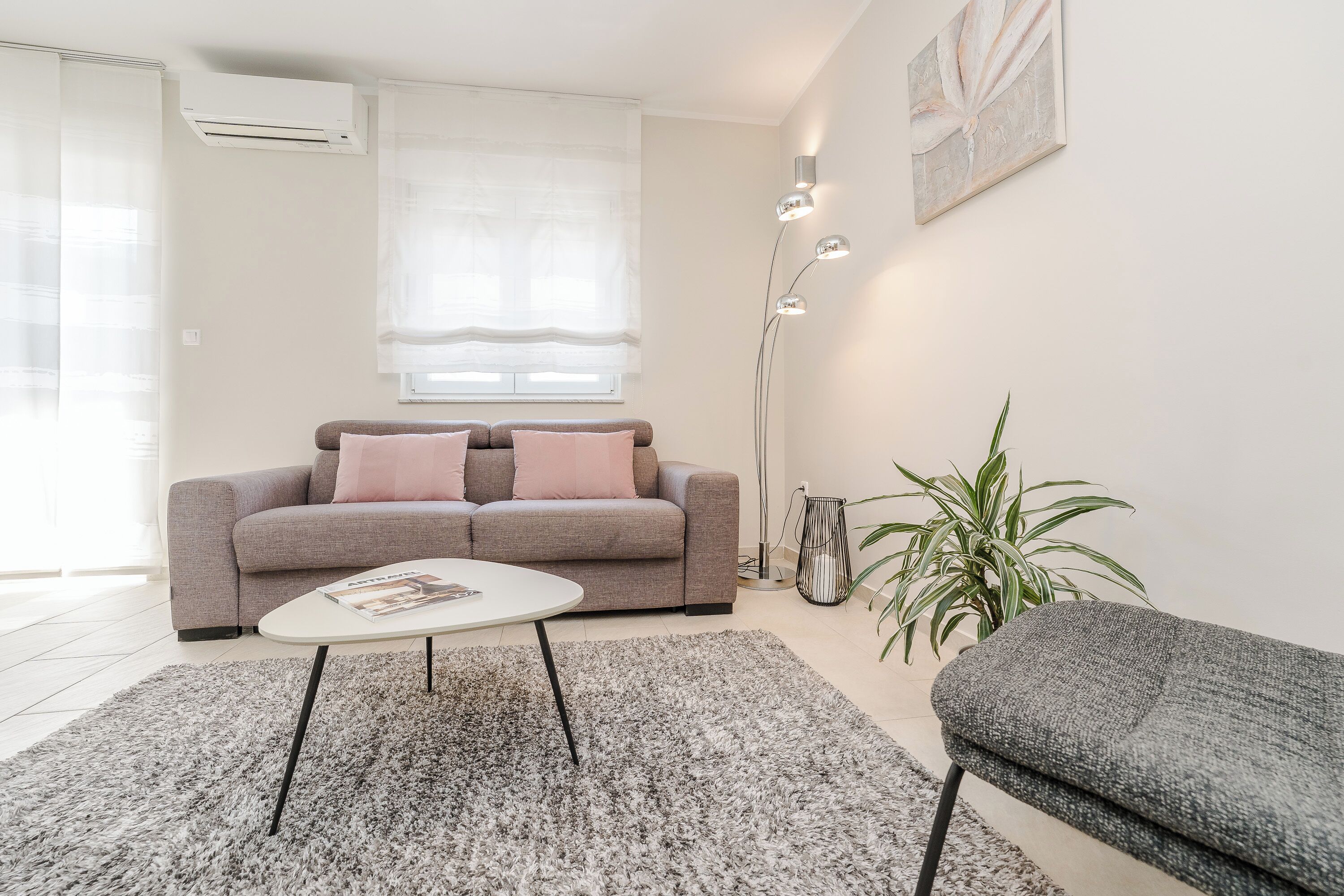 A modern living room with a grey sofa, a white coffee table, and a grey armchair. There is a floor lamp, a potted plant, and a window with sheer curtains.