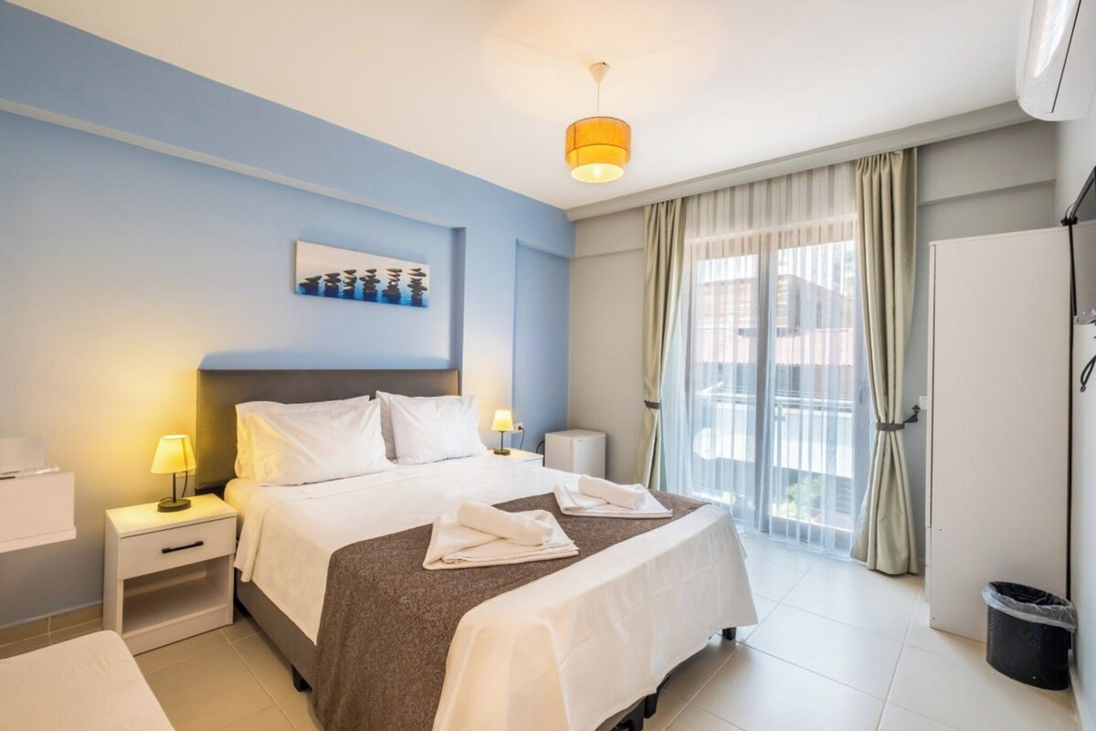 A hotel room with a bed, nightstands, a lamp, a painting, a window with curtains, and a view of the outside.