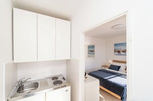 Uma cozinha compacta com pia e fogão, uma cama com manta azul, e um quadro de um farol na parede.