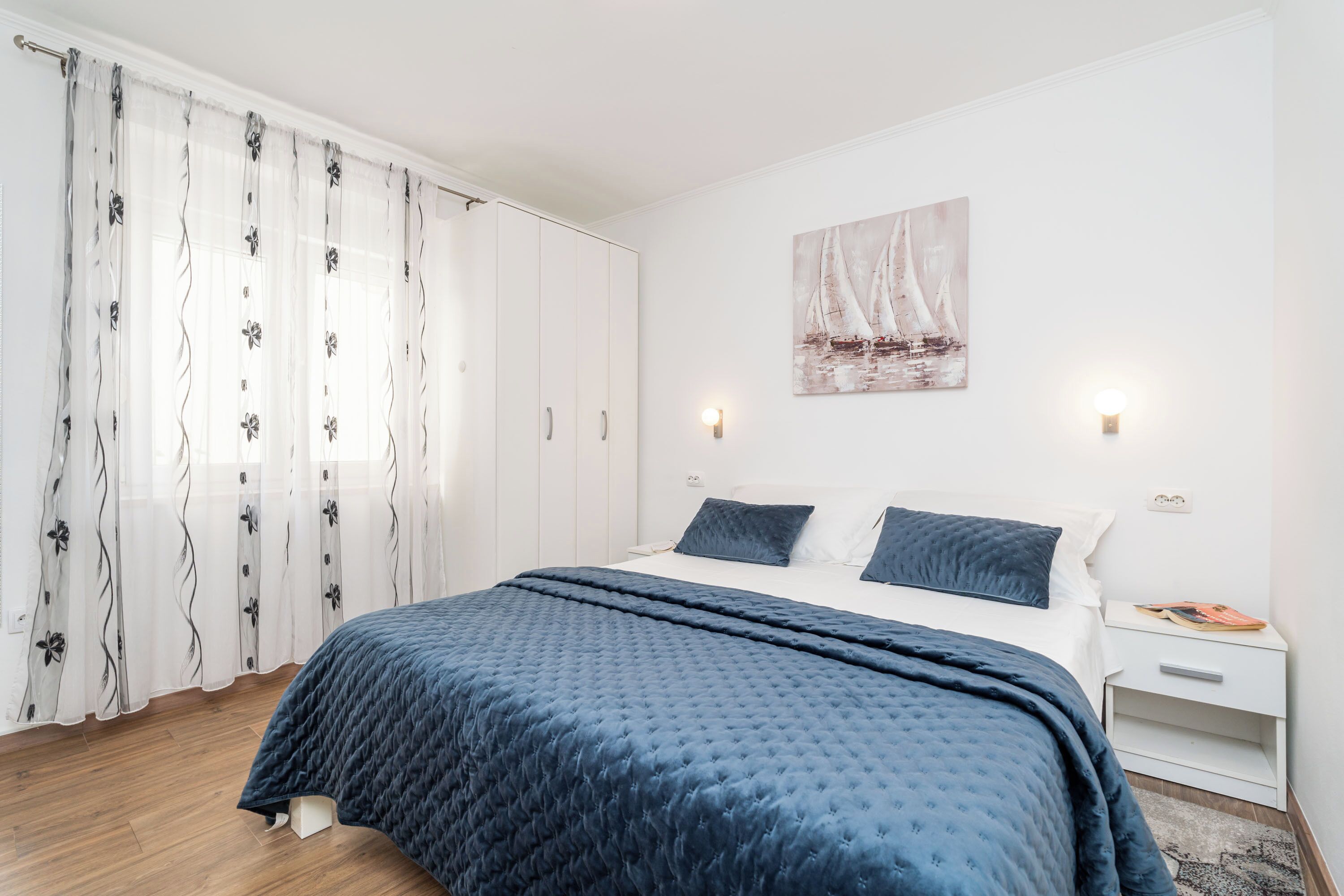Un dormitorio con cama, cortinas blancas, un cuadro y una mesita de noche.