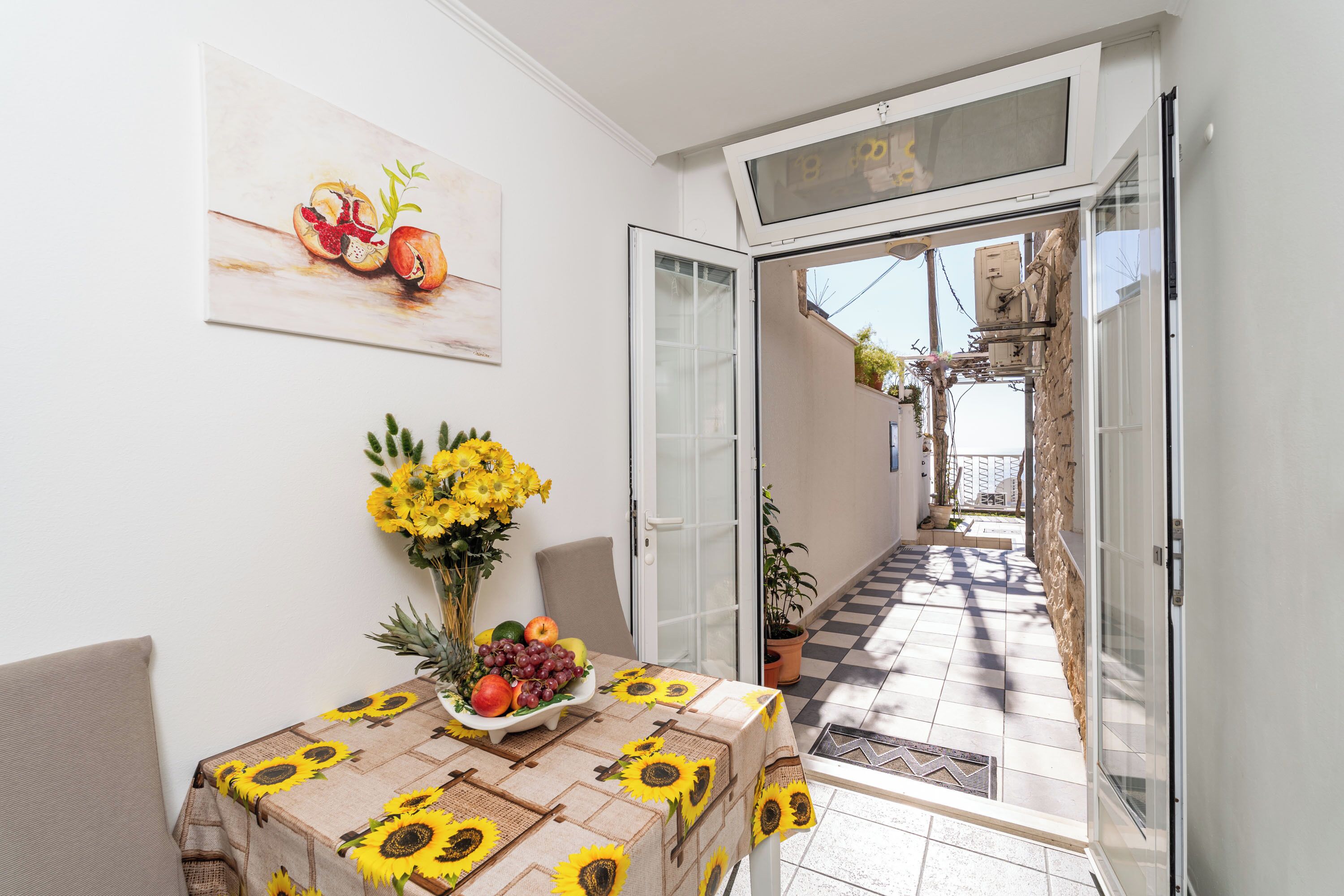 A sunlit dining area with a table set for two, featuring a sunflower tablecloth and a fruit and vegetable centerpiece.