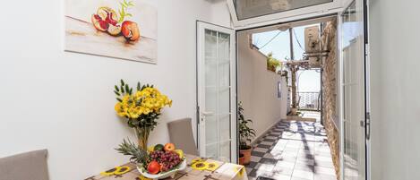 A sunlit dining area with a table set for two, featuring a sunflower tablecloth and a fruit and vegetable centerpiece.