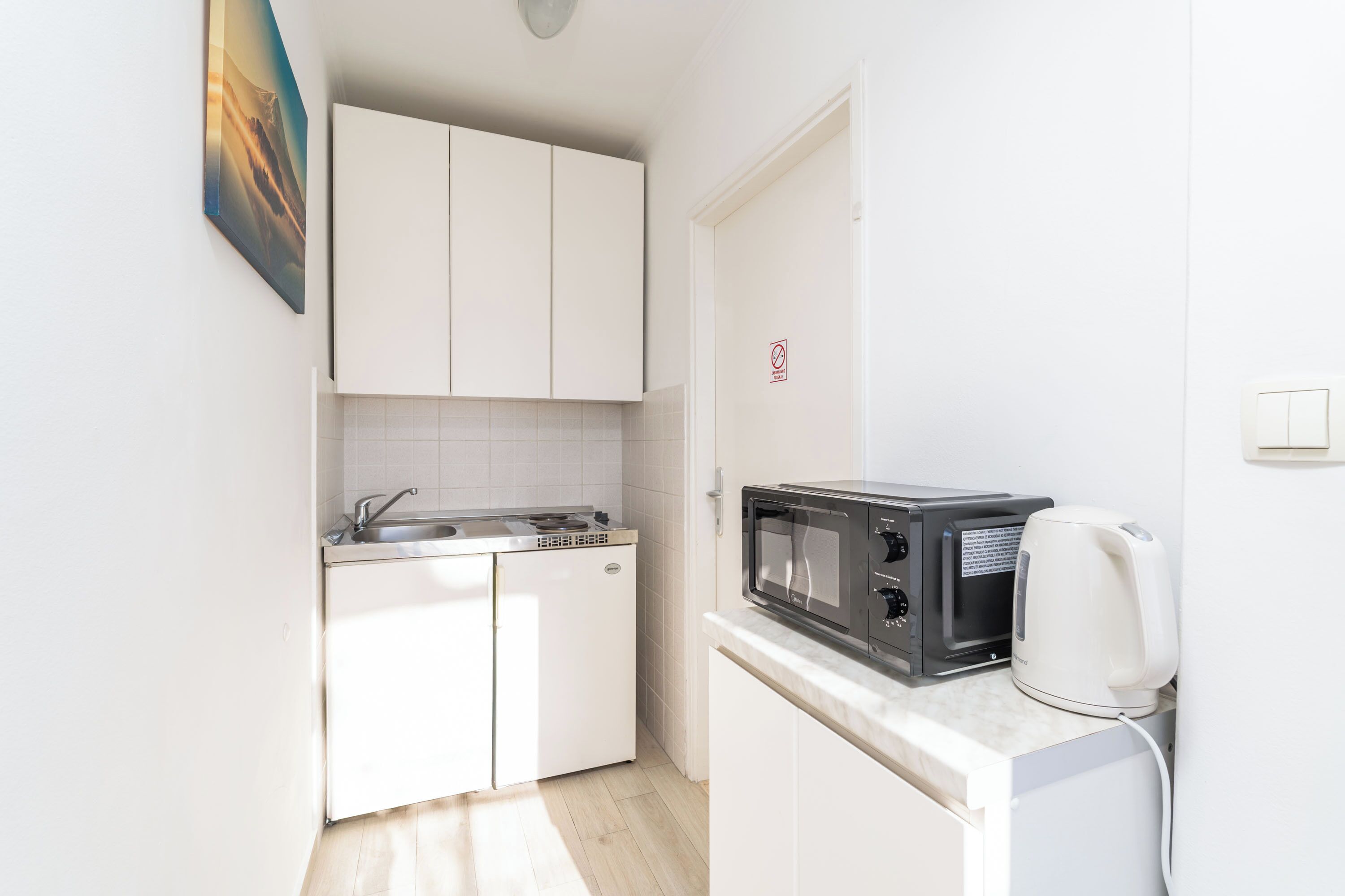 Una cocina compacta con armarios blancos, un microondas, un hervidor y un lavamanos.