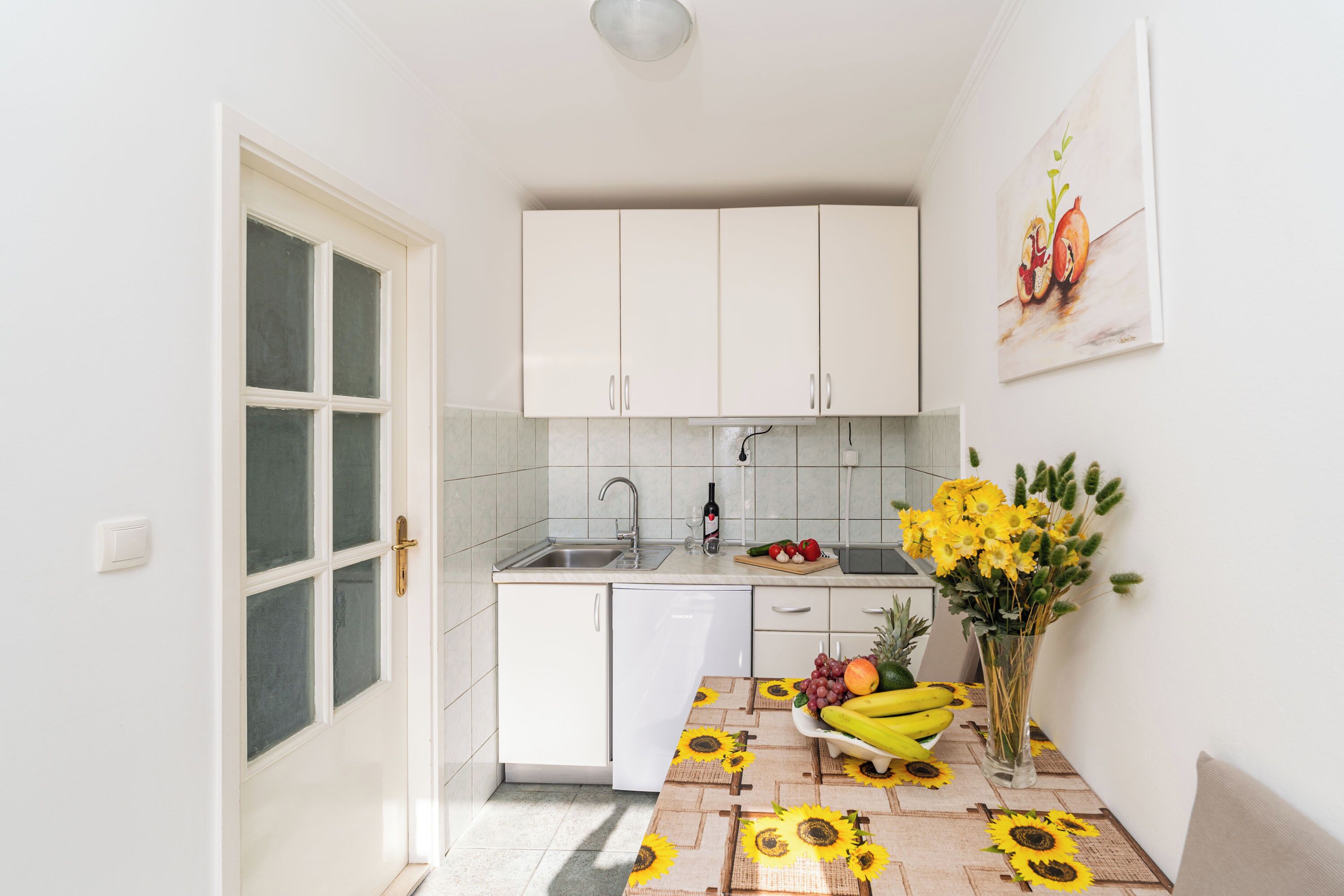 Een keuken met witte kastjes, een gootsteen en een eethoek met een tafelkleed met zonnebloemen.