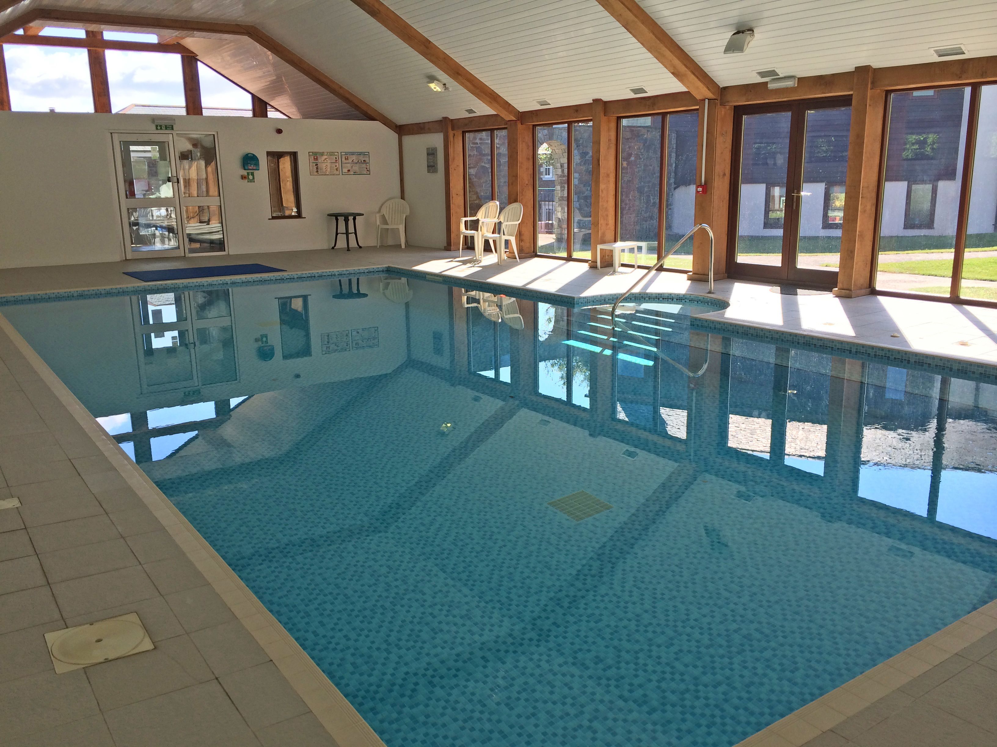An indoor swimming pool with clear blue water, surrounded by white chairs and a tiled floor.