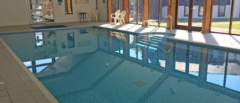 An indoor swimming pool with clear blue water, surrounded by white chairs and a tiled floor.