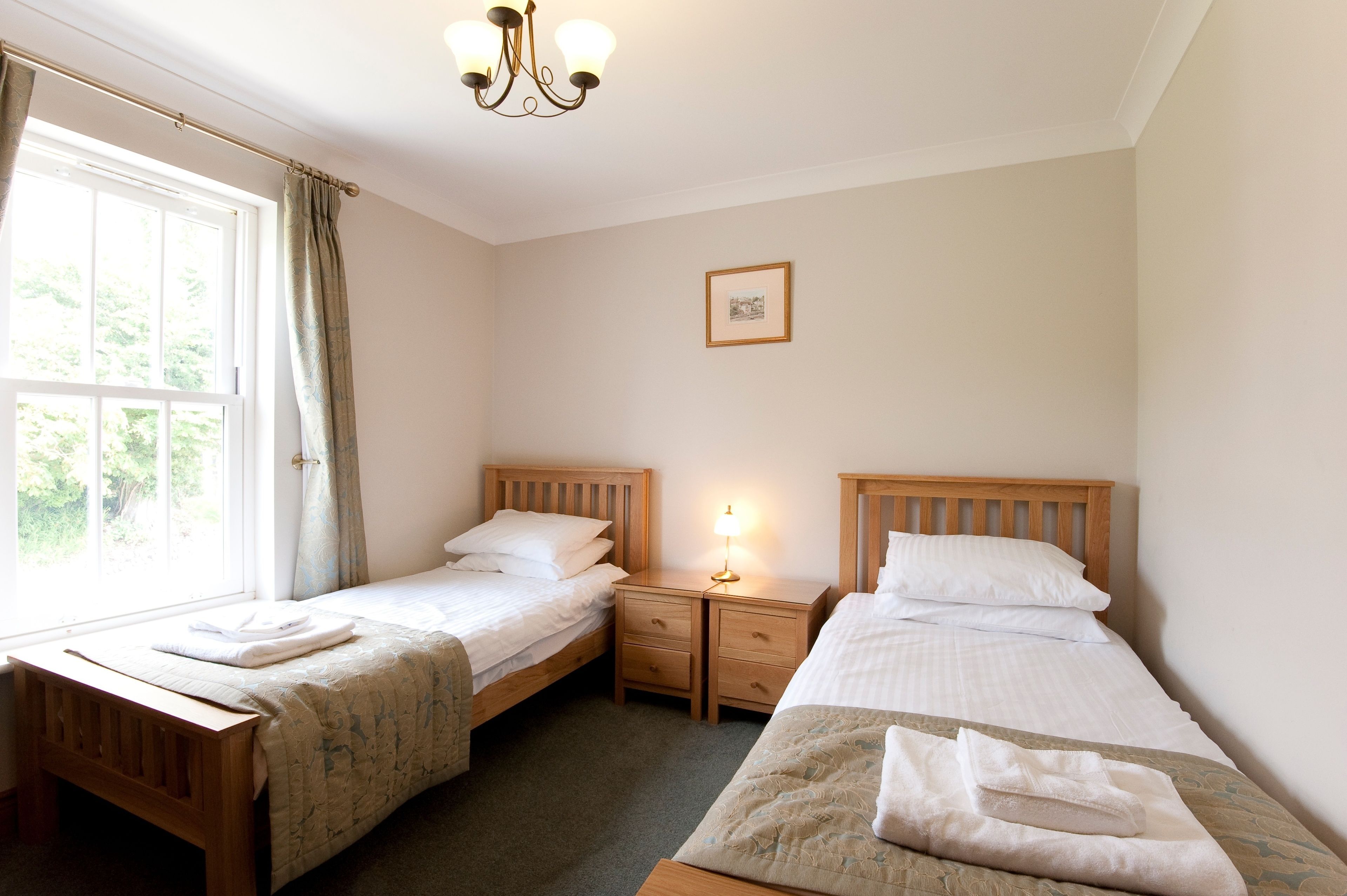Una habitación con dos camas individuales, una mesita de noche con lámpara, un cuadro en la pared y una ventana con cortinas.
