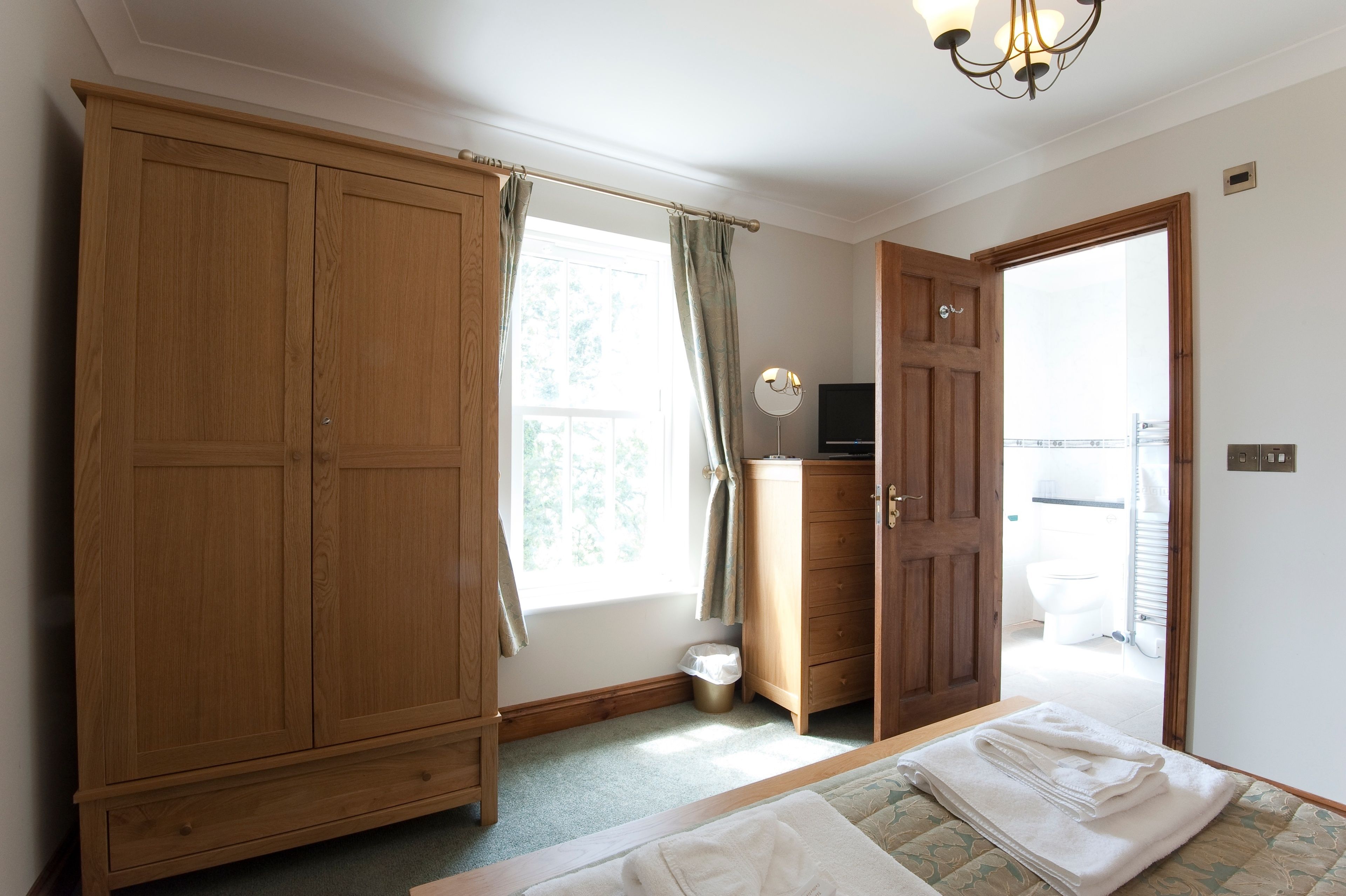 A bedroom with a large wooden wardrobe, a bedside table with a folded towel, a small cabinet, and a window with curtains.