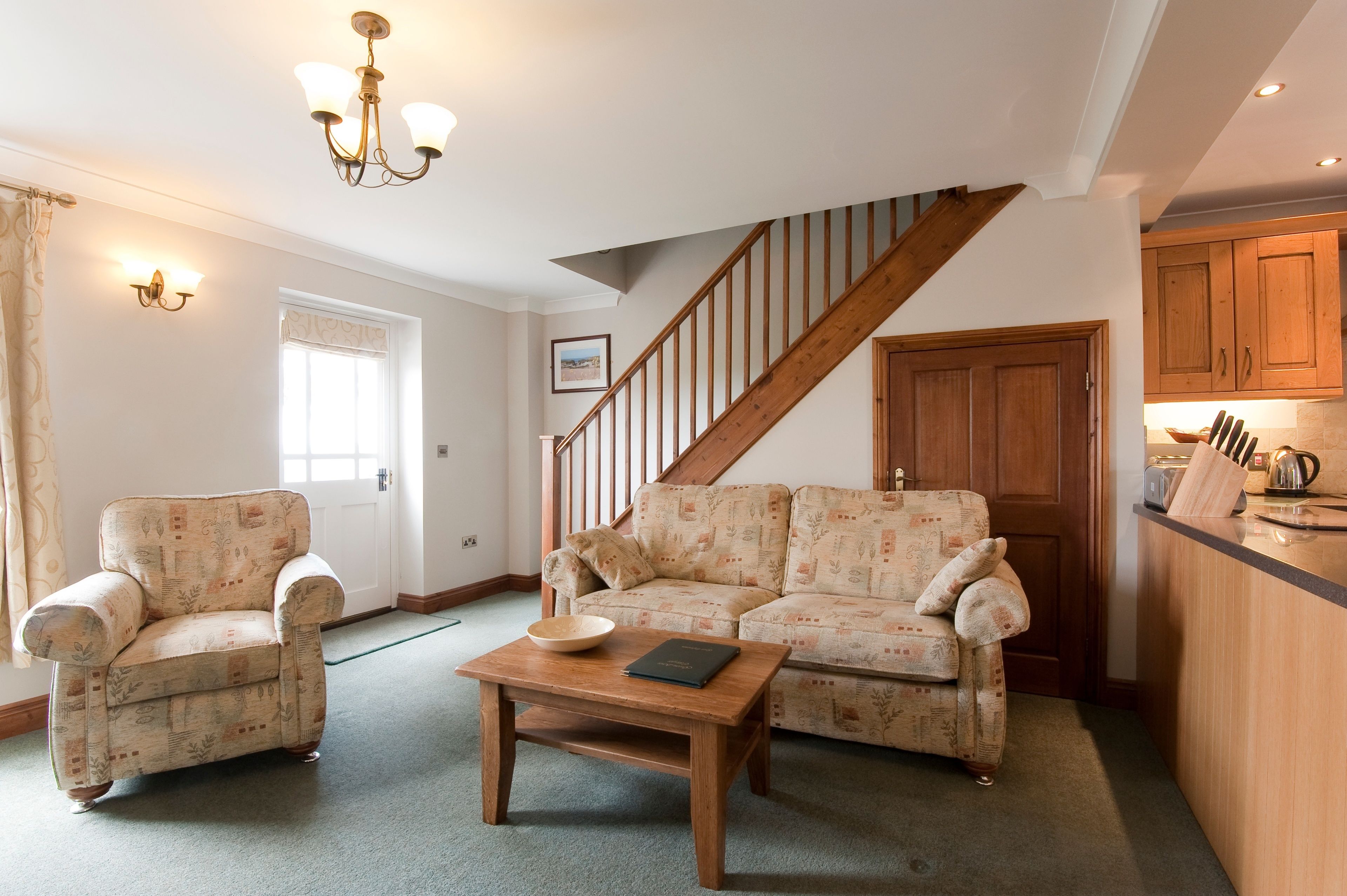 A living room with a sofa, armchair, and coffee table. A staircase leads to another room.
