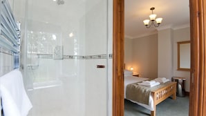 A hotel room with a glass-enclosed shower, a bed with neatly folded towels, a bedside lamp, and a wooden bench. - Green Acres Cottages (Par)