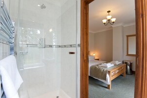 Habitación de hotel con ducha envuelta en vidrio, cama con toallas ordenadas, lámpara de noche y banco de madera.