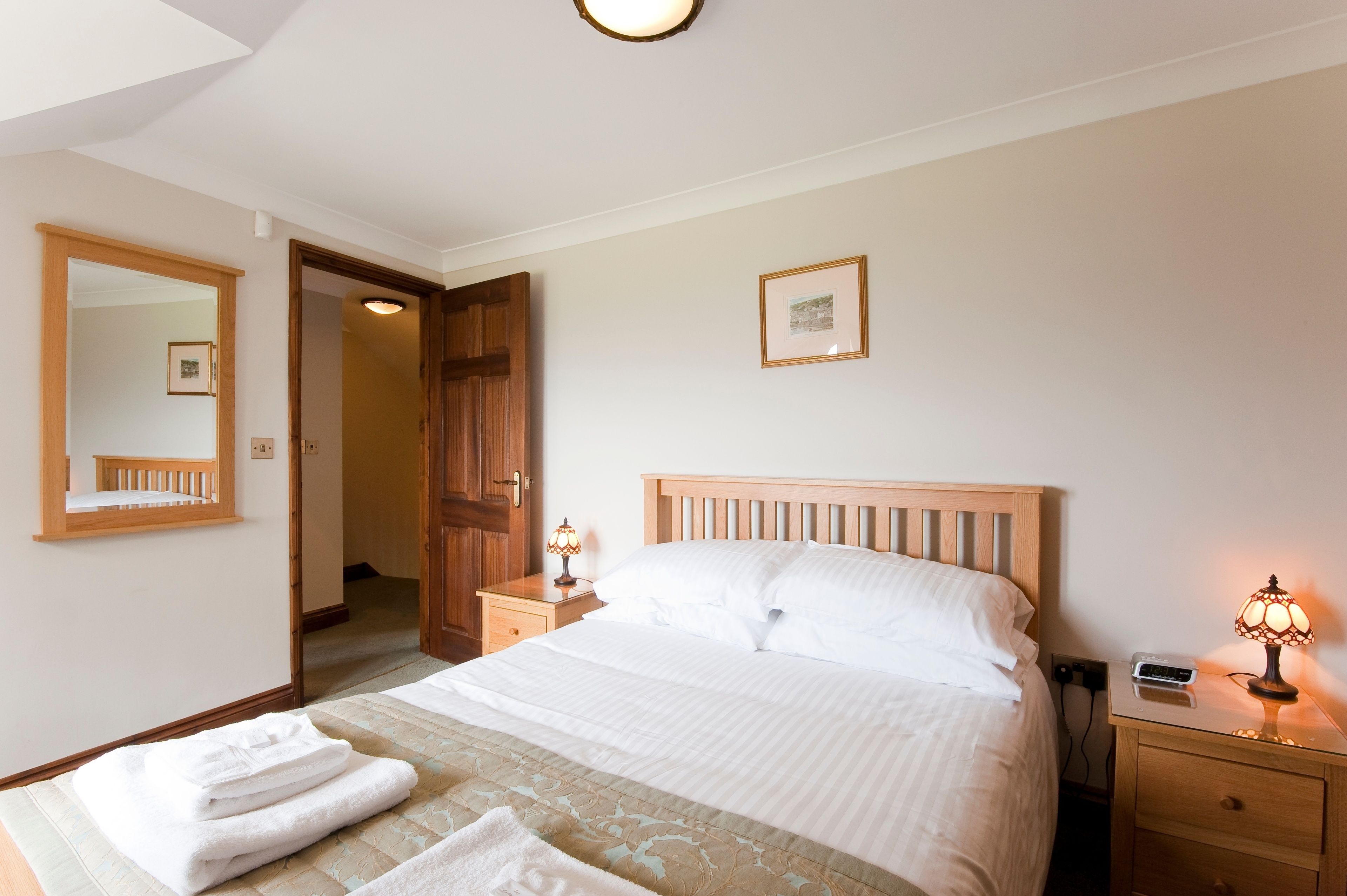 Una cama bien hecha con sábanas blancas, una lámpara de noche y una mesita de noche con lámpara. Se ve una puerta de madera y un espejo al fondo.