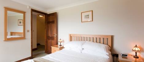 Una cama bien hecha con sábanas blancas, una lámpara de noche y una mesita de noche con lámpara. Se ve una puerta de madera y un espejo al fondo.