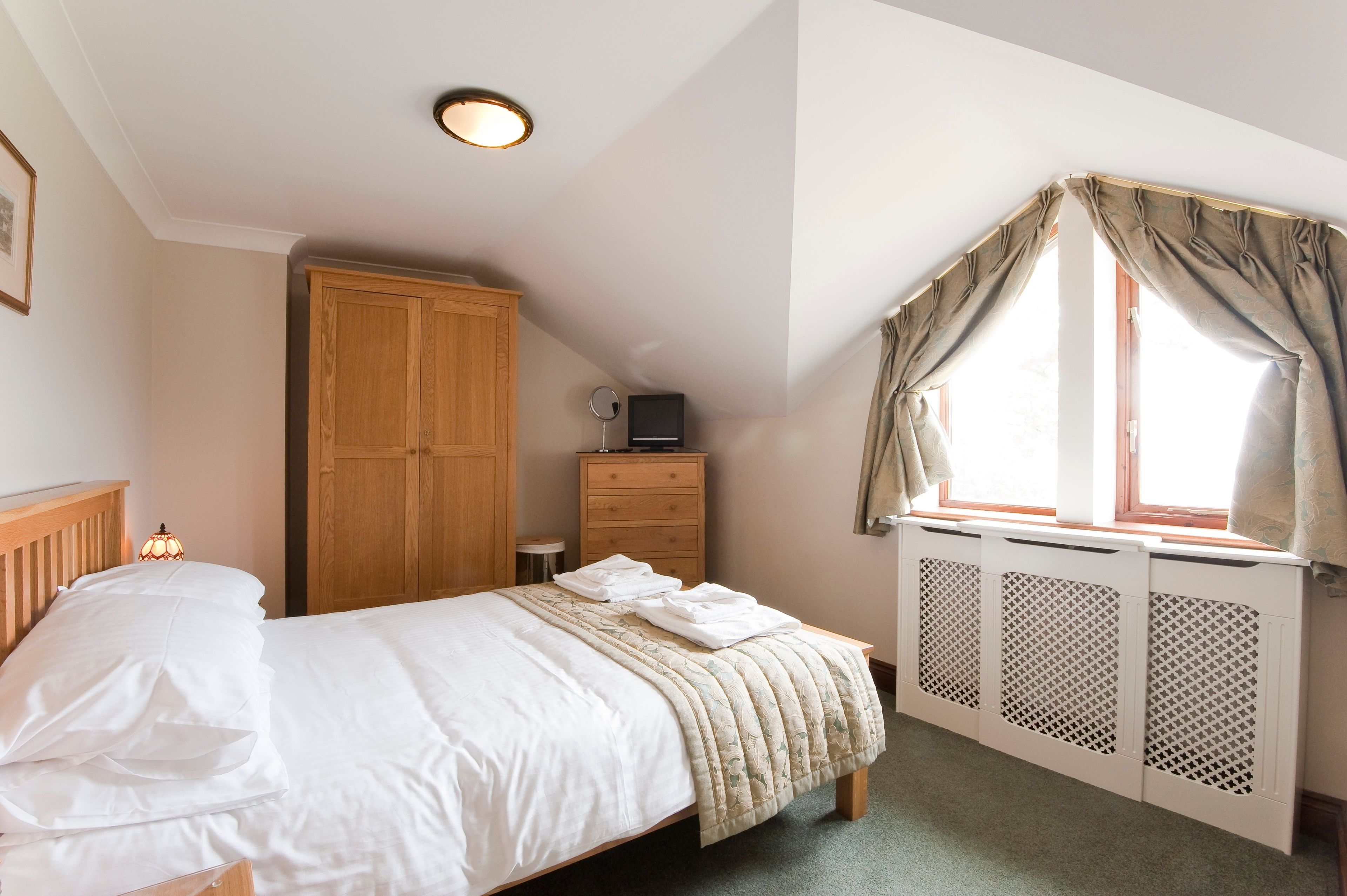 Een slaapkamer met een bed, een houten kledingkast, een klein tafeltje met een lamp en een raam met gordijnen.