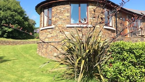 A stone building with a curved roof and large windows, surrounded by well-maintained grass and shrubs. - Green Acres Cottages (Par)